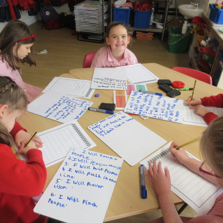 Group of young students in red uniforms sit around a classroom table, writing on whiteboards with blue markers; a smiling girl looks at the camera in the center.