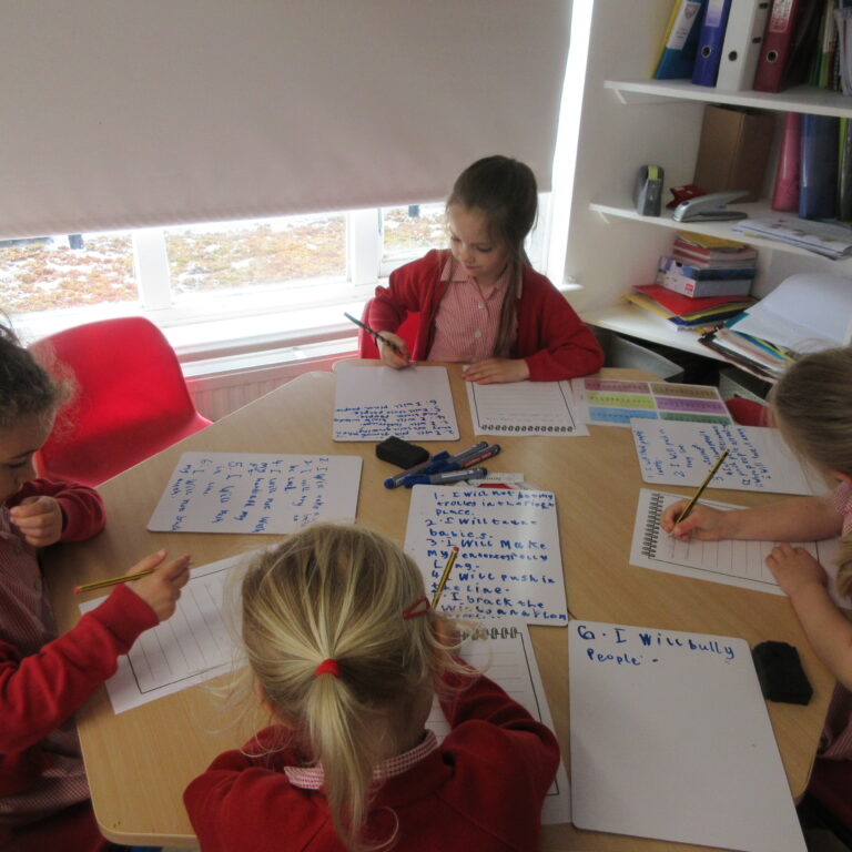 Children in red uniforms sit around a table, writing on whiteboards in a bright classroom.