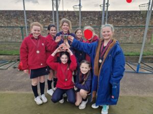 Group of young girls in red uniforms and a blue coat celebrate, holding a trophy on an outdoor sports court.