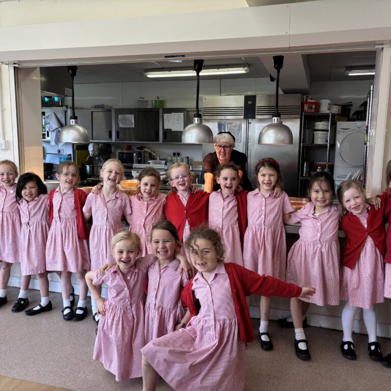 Group of smiling schoolchildren in pink check uniforms posing with a teacher in a school kitchen/cafeteria area