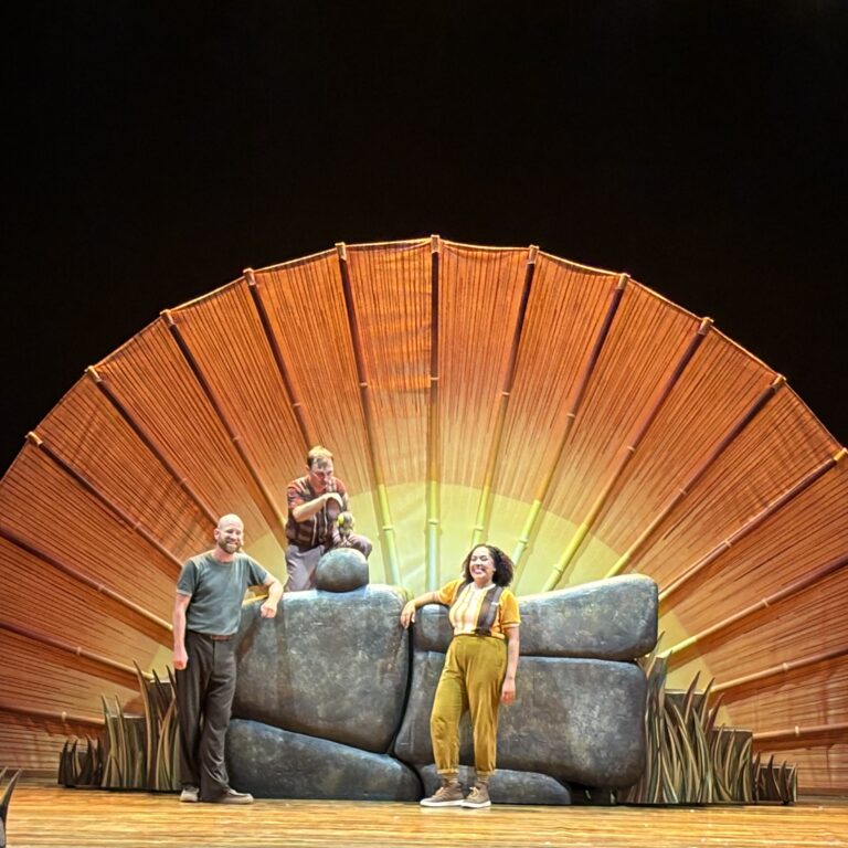 Three performers pose on a large orange fan-shaped stage prop with desert plants at the front; dark background.