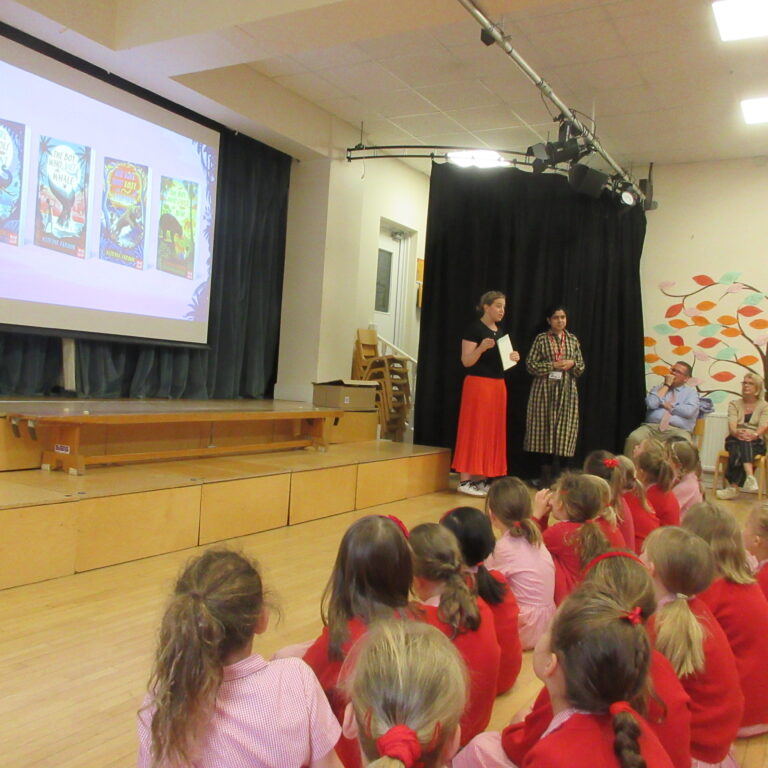 Children in red uniforms sit on the gym floor facing a stage as two presenters speak to them, with a projector screen on the left showing posters.
