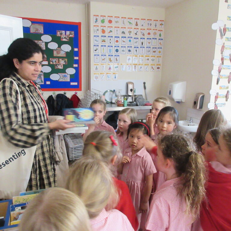 Teacher handing a book to a group of young students in a classroom; kids wearing pink uniforms, colorful classroom posters in the background.