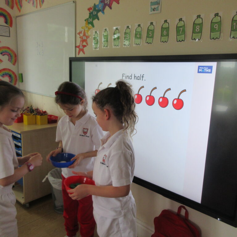Three students in white uniforms gather around a classroom activity, holding bowls with colored counters.