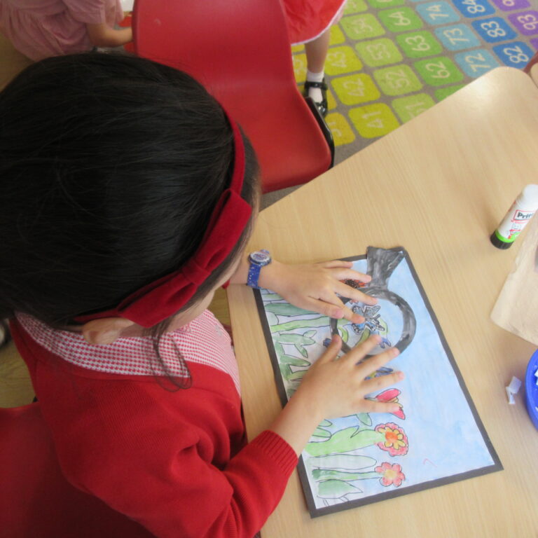 Top-down view of a child in a red shirt coloring at a wooden table with printed pictures and a blue tray of small white paper pieces nearby in a classroom.