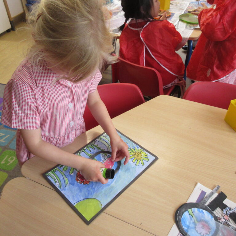 Child in a red-checkered dress sits at a table, gluing colorful paper pieces onto a drawing of sunflowers in a classroom art activity.