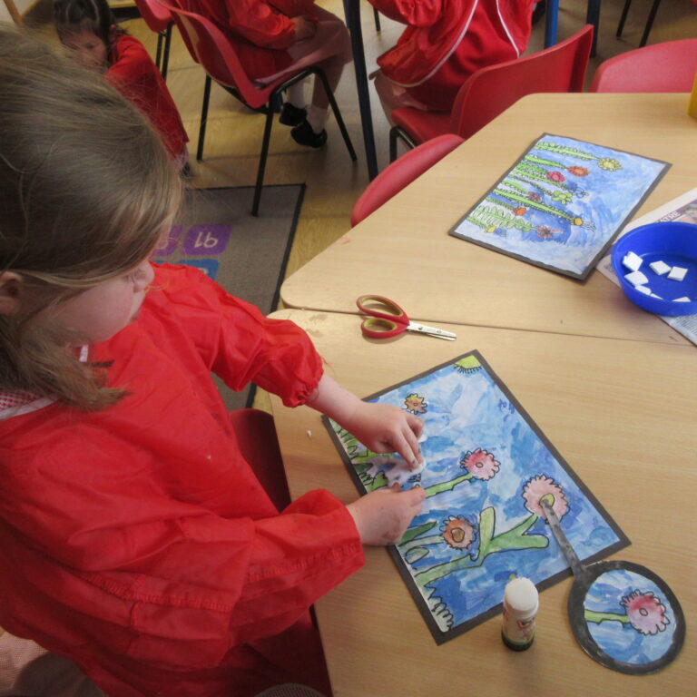 Children in red smocks seated at a table, working on colorful watercolor art projects with flower designs and blue skies on paper.