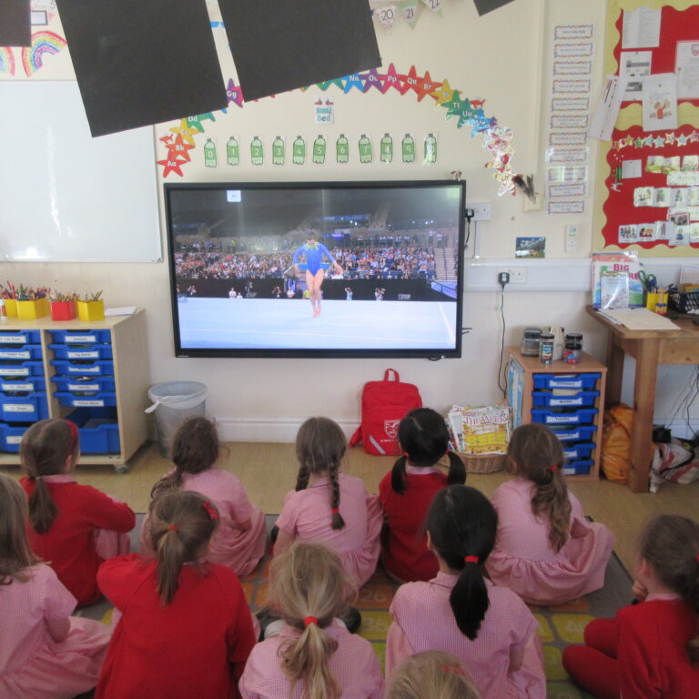 Group of young students in red and pink uniforms sit on the floor watching a gymnast on a large monitor in a classroom gym event setting.
