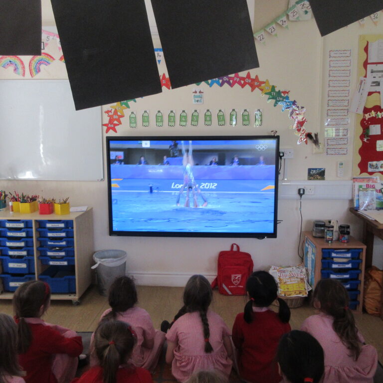 Classroom full of young students watching a gymnastics event on a large TV screen, with Olympic imagery visible on screen and colorful classroom decorations around.