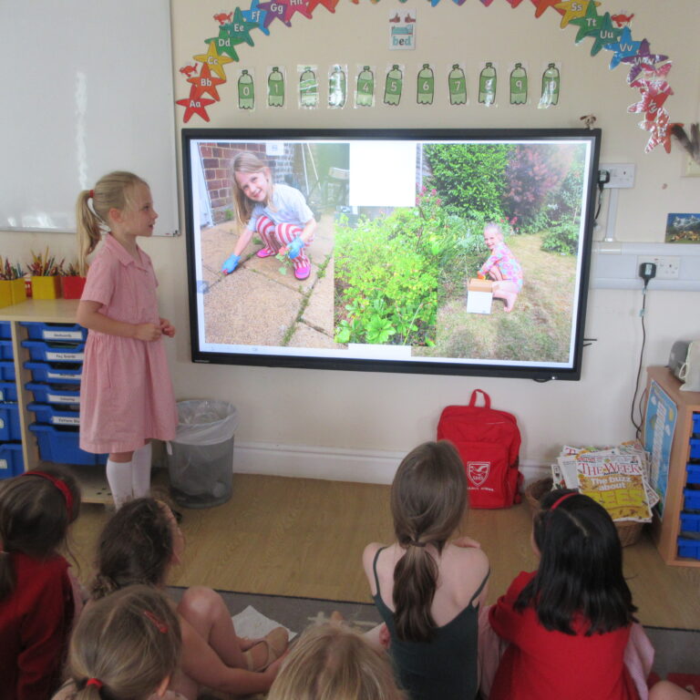 Girl in a pink dress stands beside a touchscreen displaying two outdoor garden photos as classmates watch.