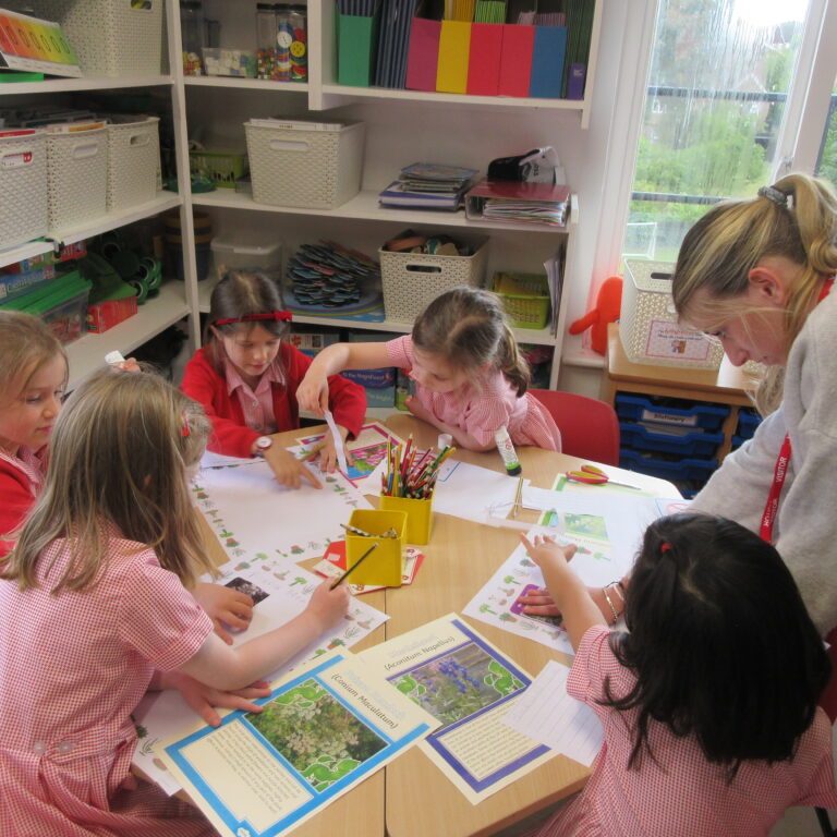 Group of young students in red checkered uniforms sitting around a table, working on craft worksheets with pencils and glue while a teacher helps on the right.
