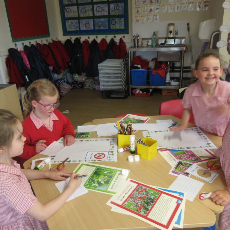 Young children in red-check uniforms sit at a table doing craft activities with worksheets and colored pencils in a classroom.