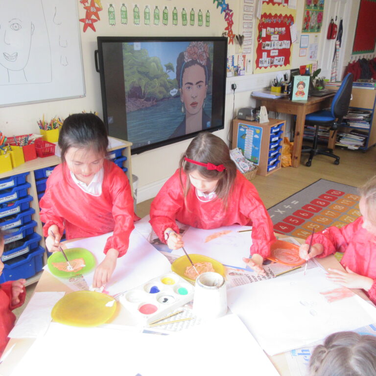 Children in red smocks painting with watercolors at a classroom art table.