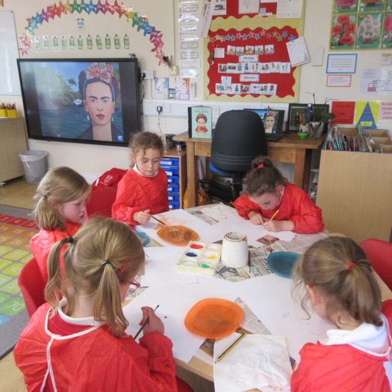Group of young students in red smocks sit around a table painting with watercolors in a bright classroom.