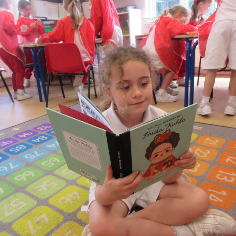 Girl sitting cross-legged on a colorful classroom rug, reading a Frida Kahlo children's book while classmates in red uniforms work at tables in the background.