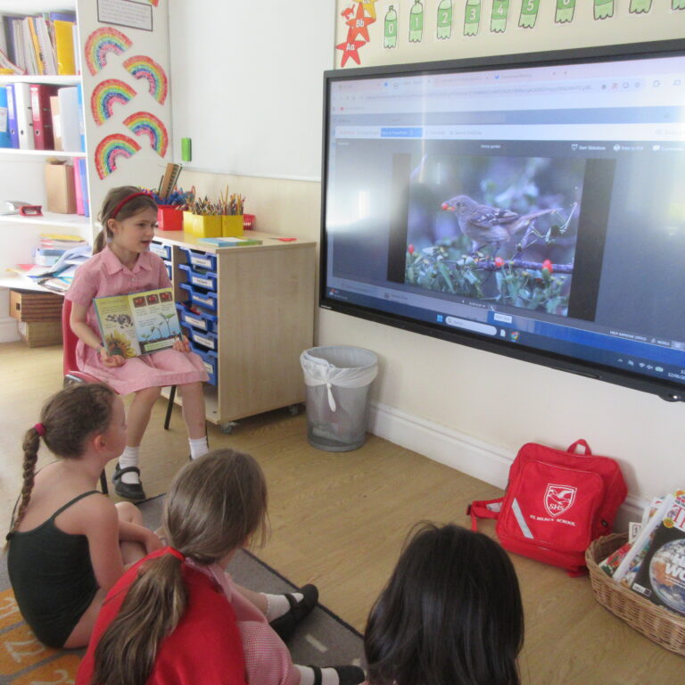 Girl in a pink uniform reads a book to seated classmates in a bright classroom; a large screen shows a bird on a branch.