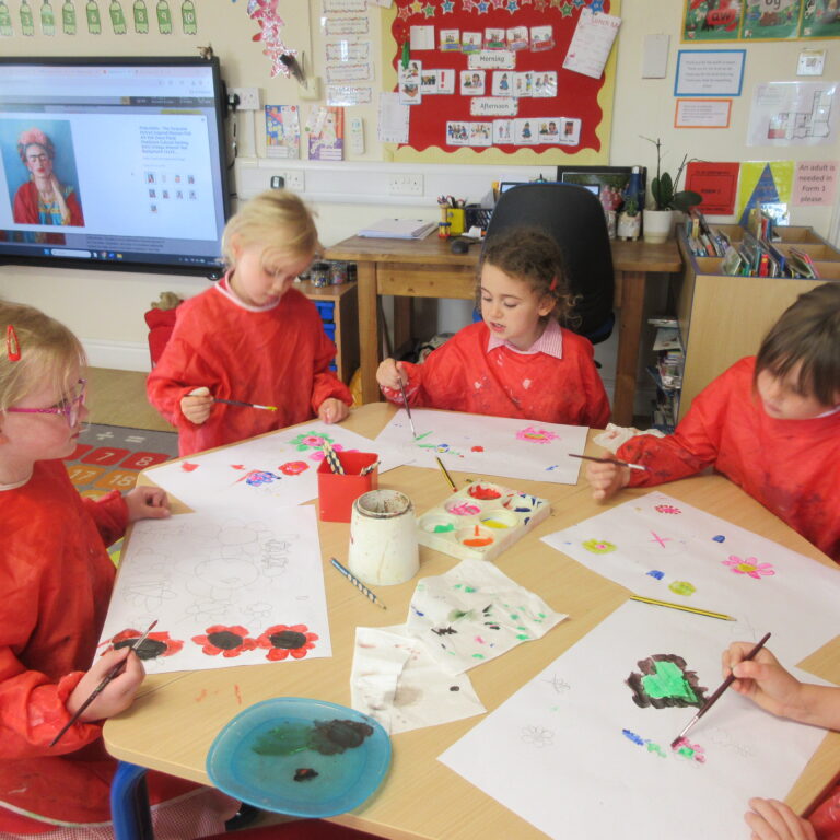 Children in red smocks painting colorful pictures at a classroom table with cups of paint and brushes.