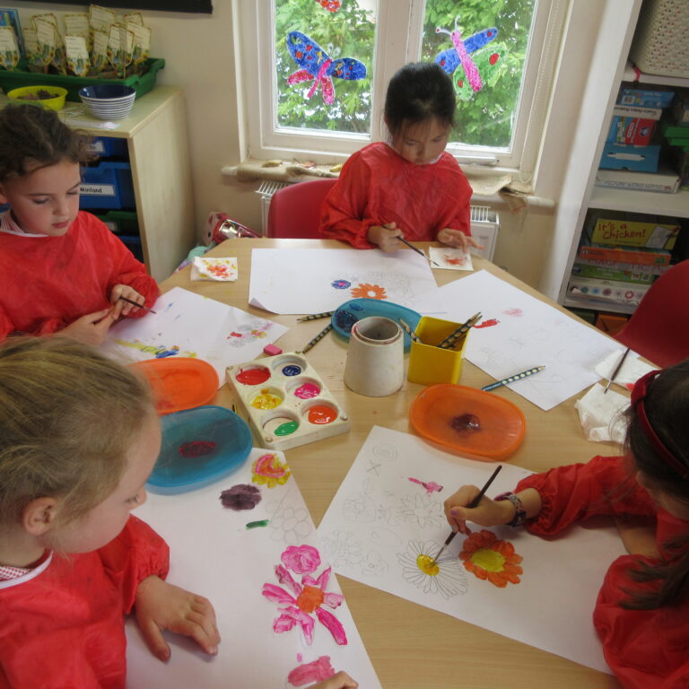 Children in red smocks paint colorful flower drawings at a classroom table.