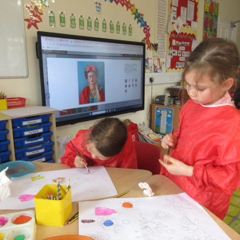 Children in red smocks painting at a classroom table; a large screen shows a Frida Kahlo portrait