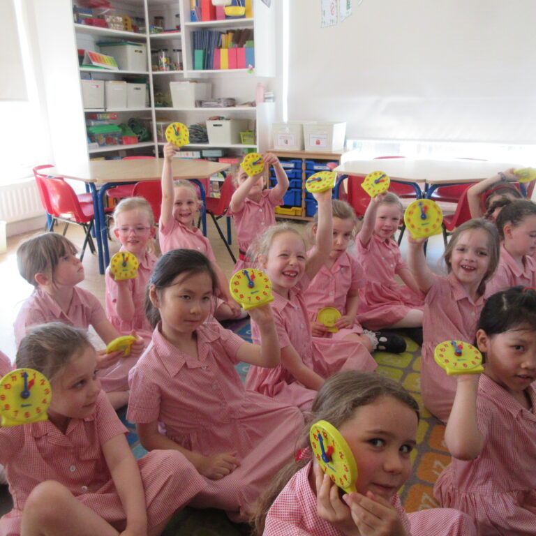 Group of smiling preschoolers in pink uniforms holding yellow toy clocks in a classroom setting.