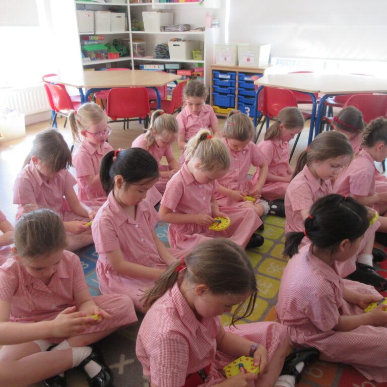 Group of young girls in pink checkered school uniforms sitting on a colorful classroom rug, each holding a yellow circular craft object and focusing on an activity in a classroom setting.