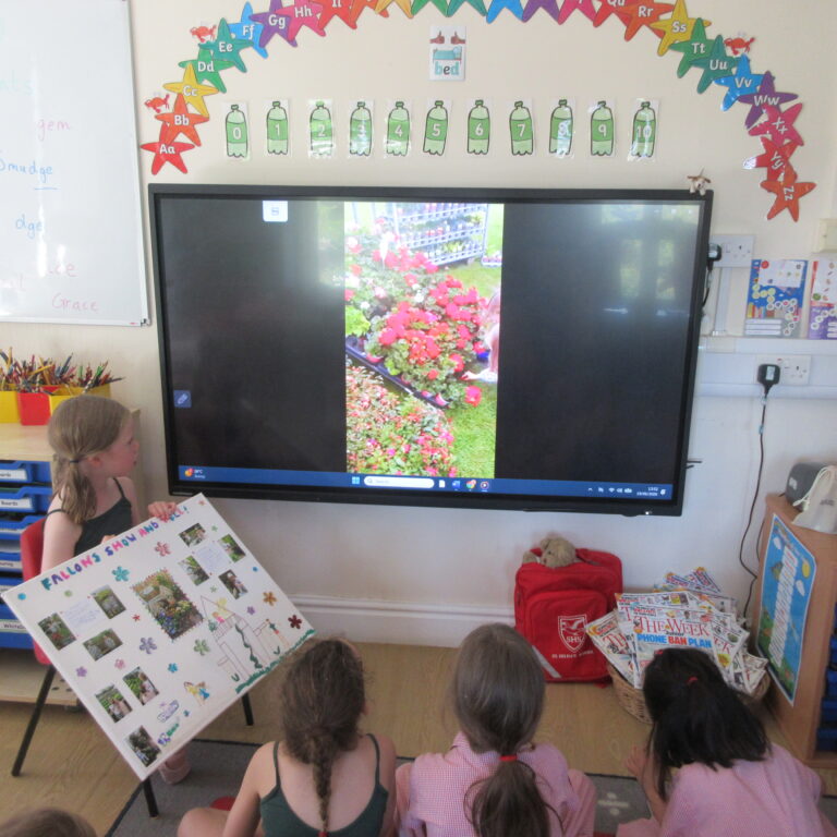 Young children sit on the floor facing a large interactive screen showing a garden photo, as a student holds a colorful poster board nearby in a classroom setting.