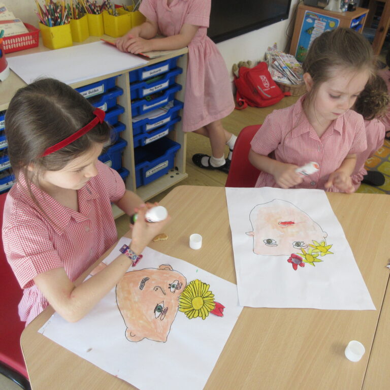 Young girl in a red-and-white check uniform colors in a classroom, drawing on a large sheet of paper at a table.