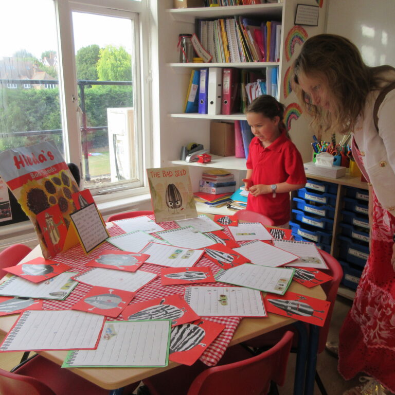 Teacher and student stand by a table covered with red cards and notebooks during a classroom craft activity.