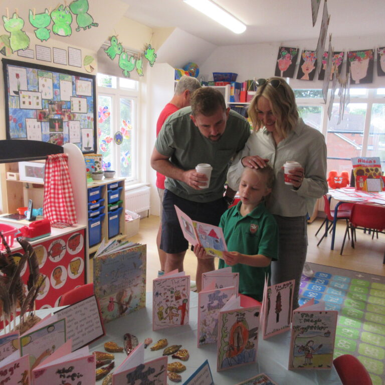 Family in a bright classroom examining a display of handmade cards and books on a table, with colorful student artwork on the walls and ceiling