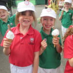 Group of primary school children in green and red uniforms eating ice cream cones outdoors, with a mural and playground in the background.