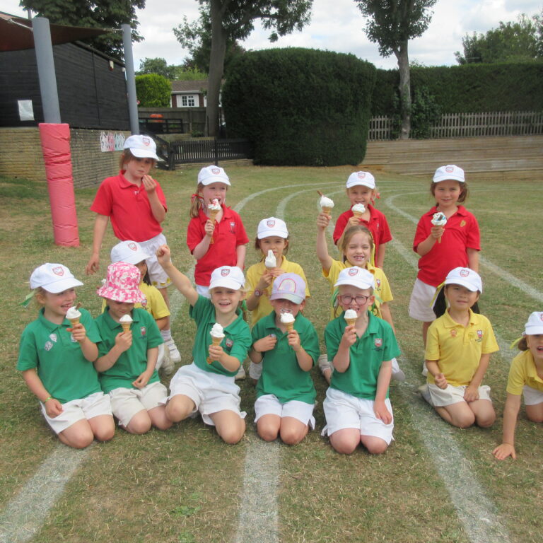 Group of schoolchildren in colored shirts kneeling on a grassy field, holding ice cream cones.