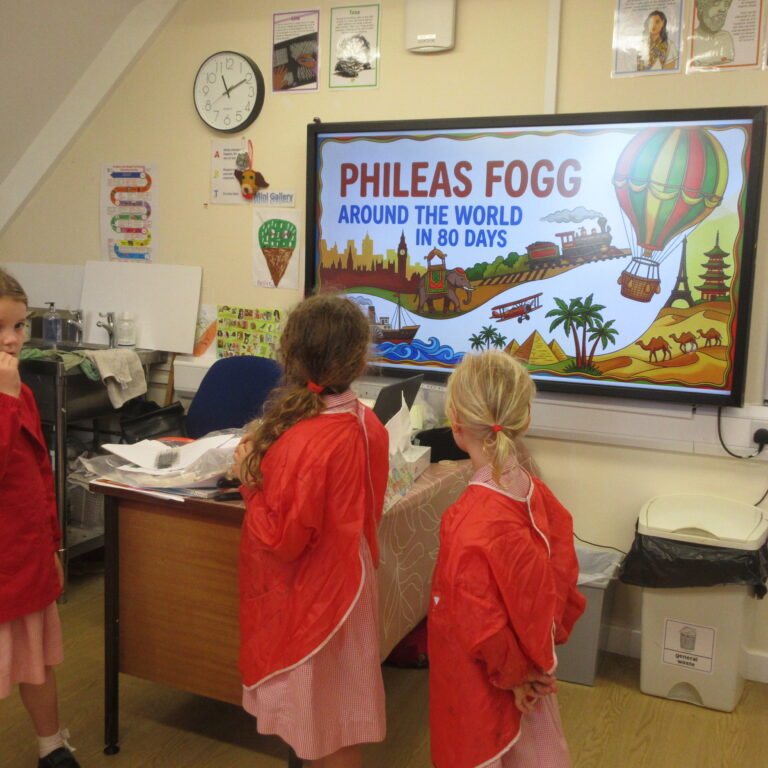 Classroom scene with children in red smocks standing around a table, viewing a large colorful poster about Phileas Fogg: Around the World in 80 Days on a screen.