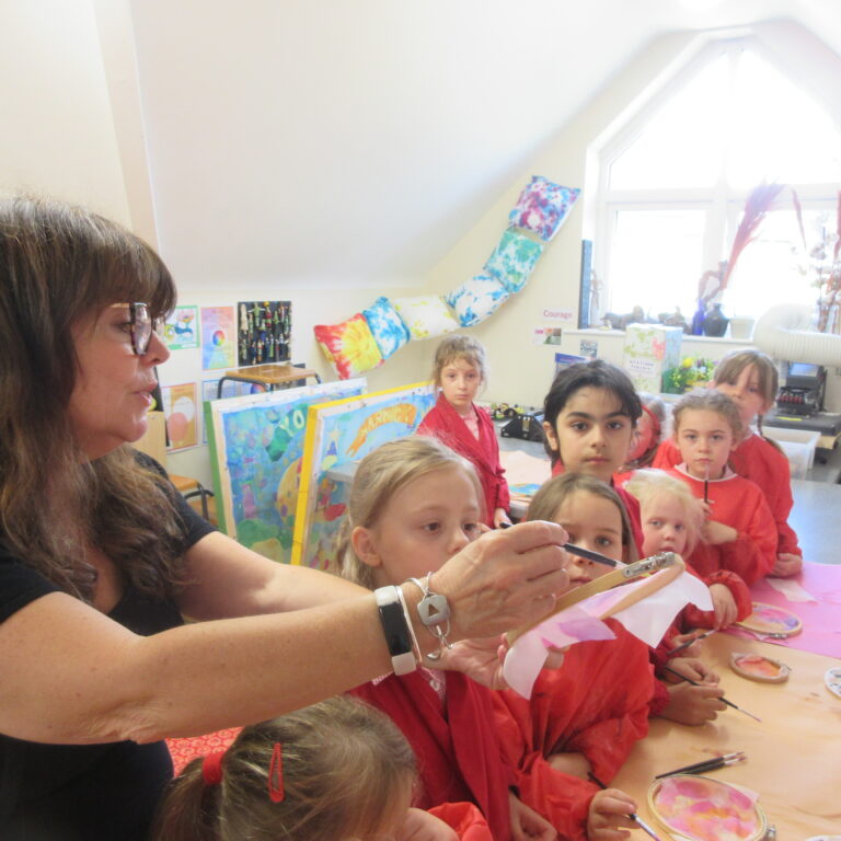 Teacher guiding a group of children painting at a table in a classroom art activity.