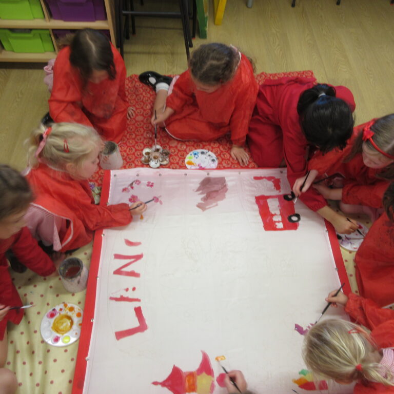 Group of children in red smocks painting a large white banner at a classroom table.
