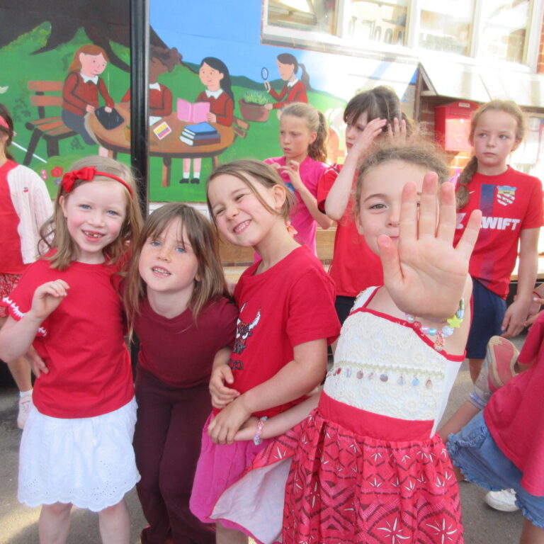 Group of smiling children in red outfits posing outdoors, with a colorful mural behind them and a brick building nearby.