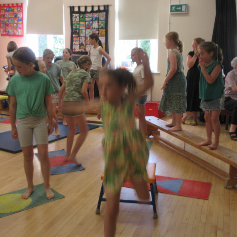 Group of children in a classroom gym balancing on low beams while adults watch nearby on benches and chairs in a bright room.