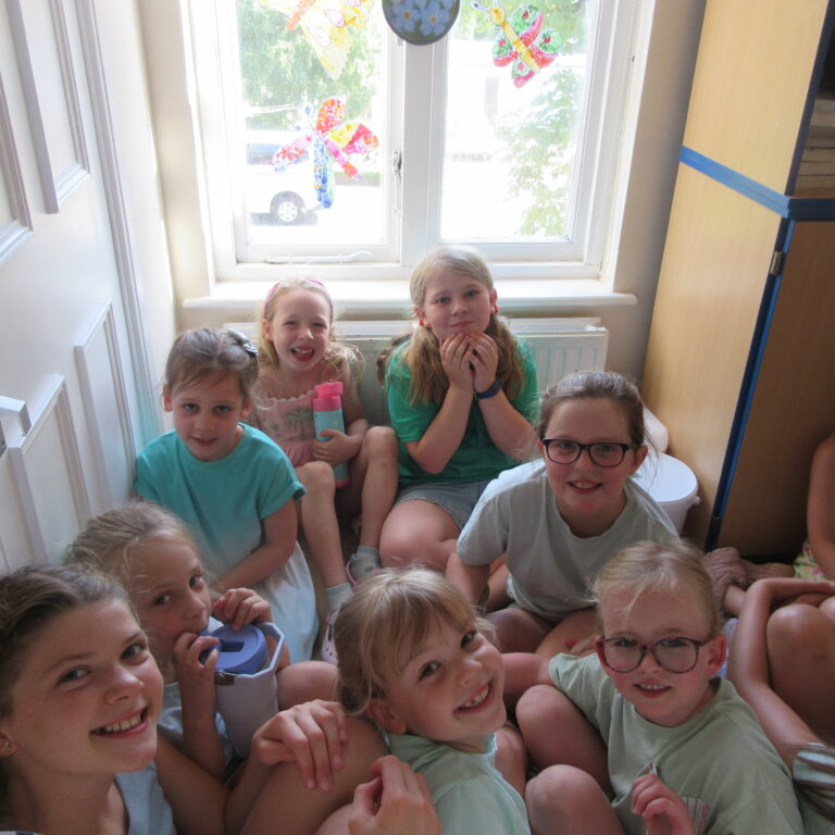 Smiling group of young children sitting on the floor in a narrow hallway by a window.