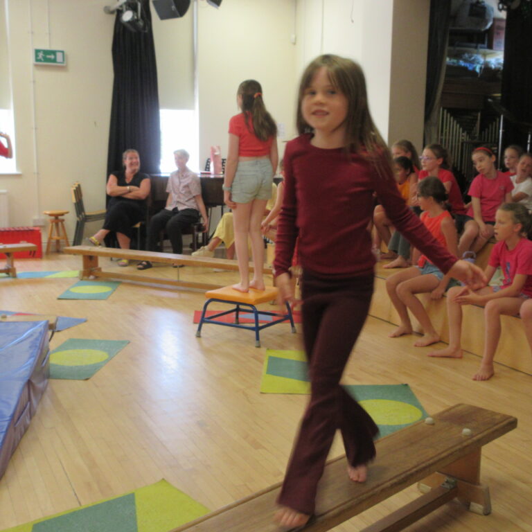 Children watching a girl in maroon long-sleeve top walk across a gym bench in a classroom or gym setting with colorful mats and equipment nearby.