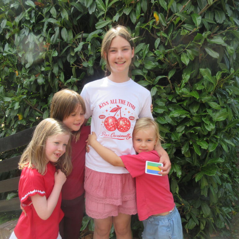 Group of smiling children posing together outdoors in front of a large green hedge; one girl in a white shirt stands center, others hug her from sides, holding a small colorful card at the bottom rightbed of the image.