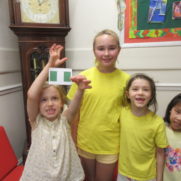 Group of smiling children indoors; a girl in a yellow shirt holds up a green-and-white card toward the camera, others around her.