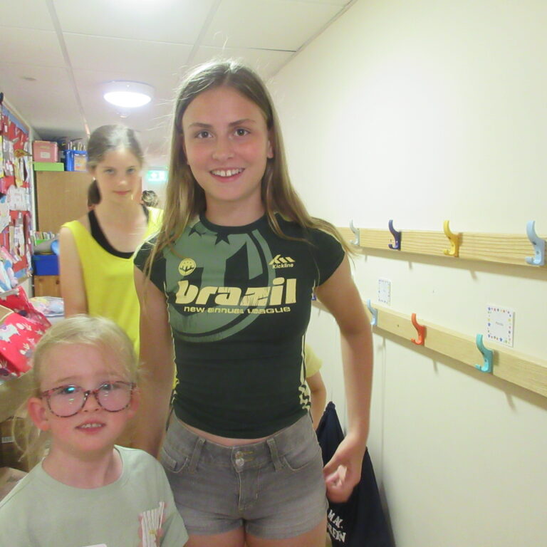Front-facing teenage girl smiles at the camera in a hallway, with another girl in a yellow top in the background and coats on hooks to the right.