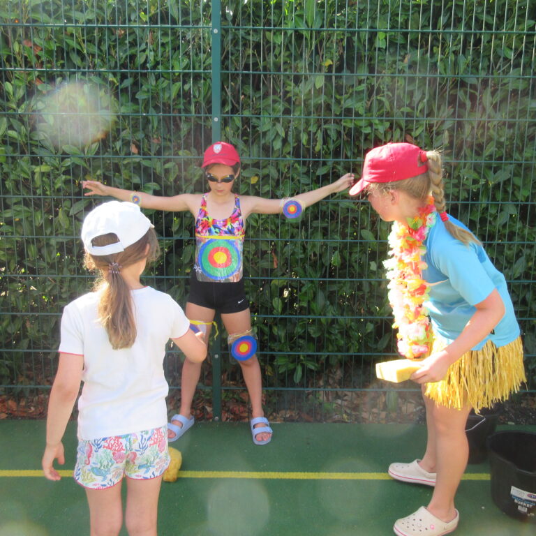 Children in red caps and colorful outfits play outdoors behind a green fence, displaying circular target stickers on their arms and legs.