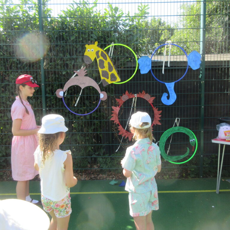 Children gather at an outdoor craft booth with colorful animal shapes hanging on a green fence behind them.