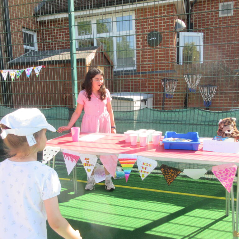 Girl in a pink dress standing behind a red table with cups at a small outdoor party by a wire fence and brick building. Flags decorate the fence behind her.
