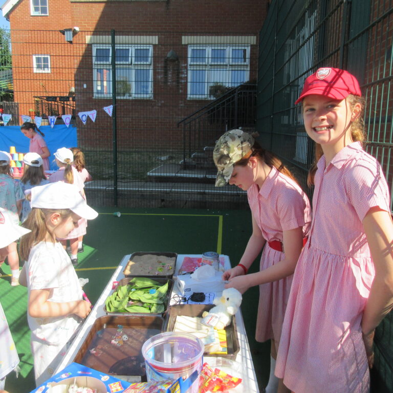 Group of young children in pink uniforms at an outdoor school event, gathered around a craft table with trays and colorful materials in front of a brick school building.