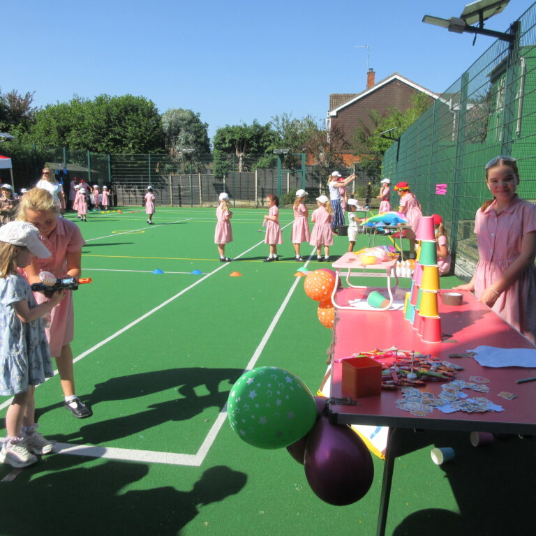 Group of young girls in pink dresses on a bright green sports court, with party tables, balloons and a fence in the background.