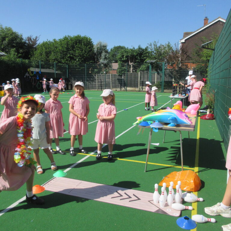 Group of girls in pink dresses at an outdoor school event on a green court with bowling pins and inflatable toys nearby.