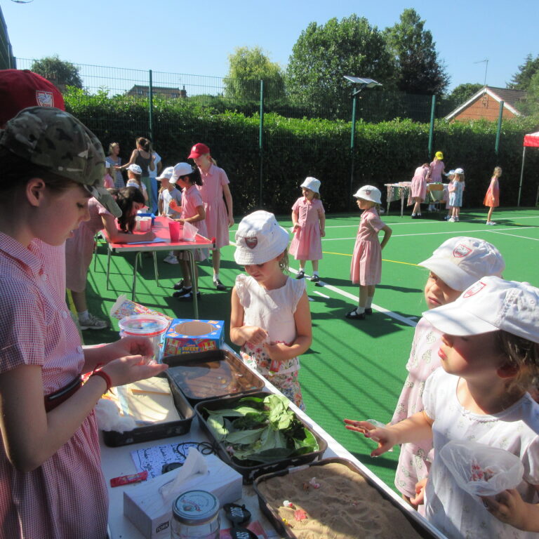 Group of young children in pink dresses and hats gathered around tables, making crafts at an outdoor event on a green sports court under a sunny sky.