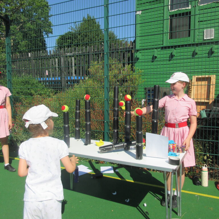 Young girls in pink dresses and white hats stand around a game table outdoors behind a green metal fence on a sunny day, with tennis balls and black tube targets set up on the table.
